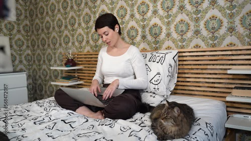 Beautiful woman is working on the computer while listening to music on headphones. The woman is sitting on the bed and petting her cat.