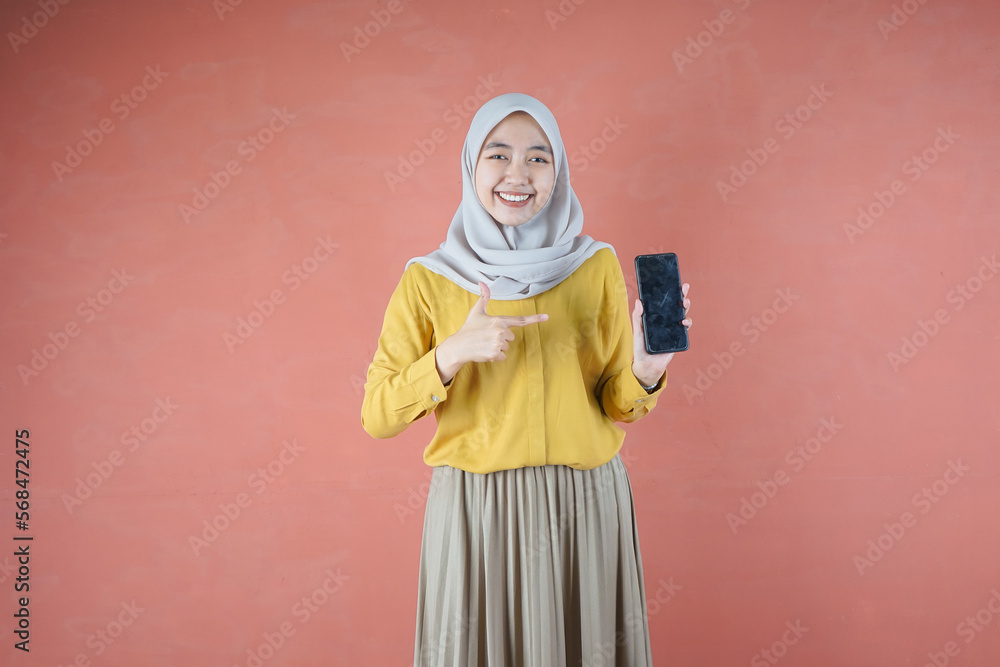 Fototapeta premium Beautiful Asian woman in yellow shirt and hijab smiling cheerful has smartphone with black screen in hand, pointing with index finger at product on brown background