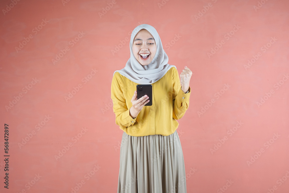 Beautiful Asian woman in yellow shirt and hijab smiling cheerful using mobile phone gesturing yes with raised fist on brown background Beautiful Asian woman in yellow shirt and hijab smiling cheerful using mobile phone gesturing yes with raised fist on brown background