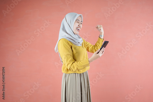 Beautiful Asian woman in yellow shirt and hijab smiling cheerful using mobile phone gesturing yes with raised fist on brown background