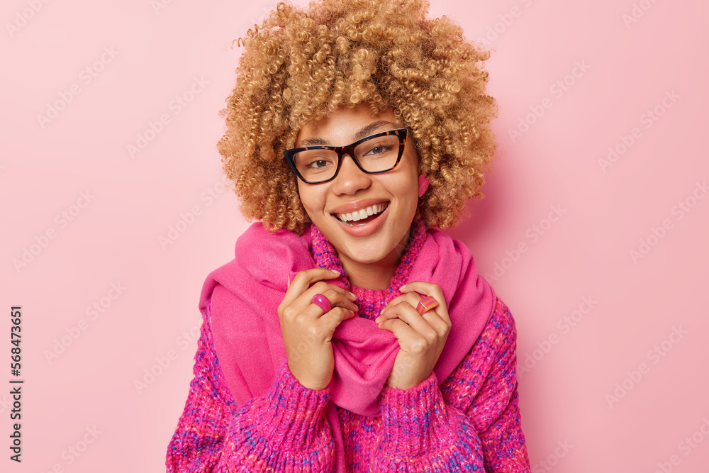 Portrait of lovely curly haired woman smiles broadly shows white teeth wears pink knitted jumper and scarf around neck looks through spectacles isolated over pink background. Happy emotions concept Portrait of lovely curly haired woman smiles broadly shows white teeth wears pink knitted jumper and scarf around neck looks through spectacles isolated over pink background. Happy emotions concept