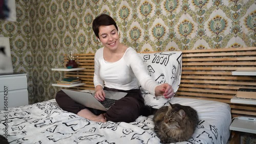 Beautiful woman is working on the computer while listening to music on headphones. The woman is sitting on the bed and petting her cat.