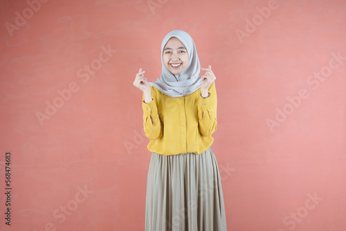 Beautiful Asian woman in yellow shirt and hijab smiling cheerfully showing Korean heart with two fingers crossed, express joy and positivity over brown background © randiwicaksono