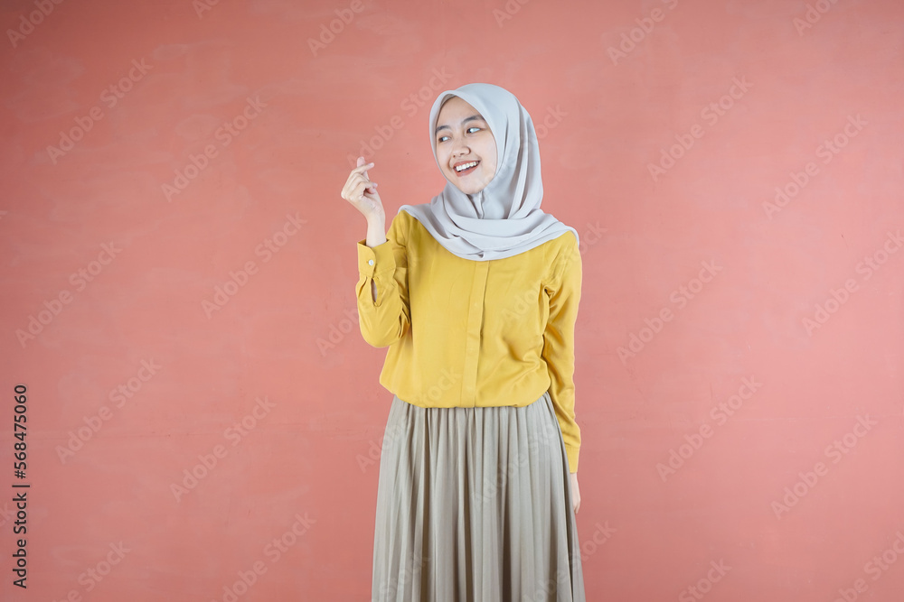 Beautiful Asian woman in yellow shirt and hijab smiling cheerfully showing Korean heart with two fingers crossed, express joy and positivity over brown background