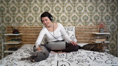 Beautiful woman is working on the computer while listening to music on headphones. The woman is sitting on the bed and petting her cat.