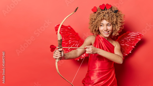 Horizontal shot of cheerful female cupid shoots arrow targets on something wears red dress with wings behind back looks mysteriously somewhere isolated over red background copy space for your text photo