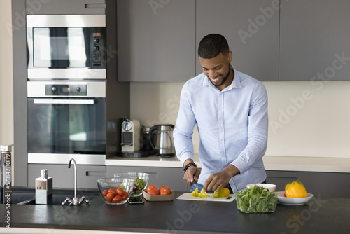 Happy young African chef man enjoying cooking hobby in contemporary home kitchen, chopping fresh vegetables on boars, preparing salad, smiling, laughing with appliance in background
