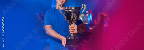 Concept banner cyber gaming esport. Winner gamer young man holding cup trophy prize of victors online video games tournament. Soft focus  neon color