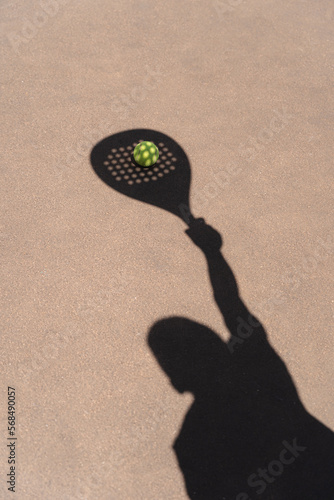 High angle of yellow ball placed on court and covered with shadow of crop person holding padel racket