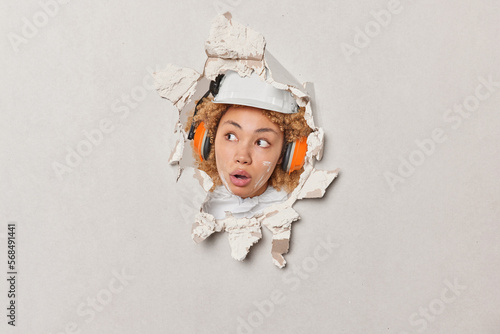 Stunned woman wears protective helmet and protection headphones looks wondered aside cannot believe own eyes breaks through plaster wall isolated over white background. People and building concept photo