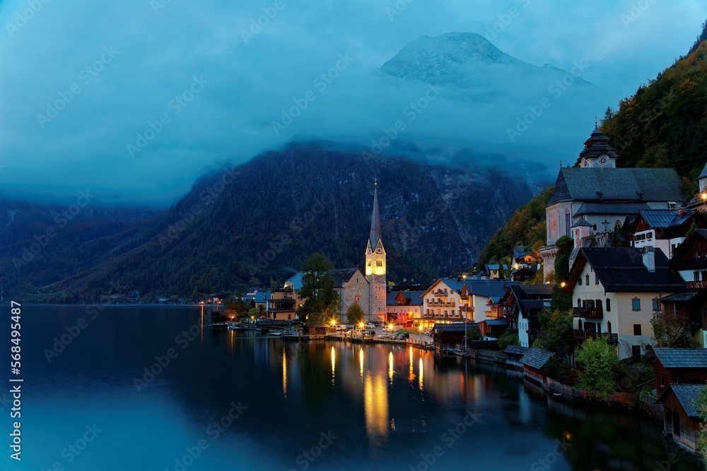 View of Hallstatt before dawn with foggy alpine mountains in background & lights of the church & town houses reflected on lake water in twilight, a peaceful lakeside village in Salzkammergut, Austria