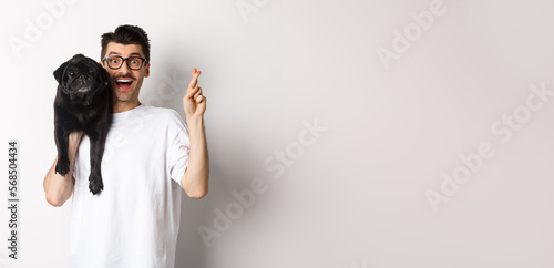 Hopeful smiling dog owner making a wish, holding cute black pug on shoulder and cross fingers for good luck, white background photo