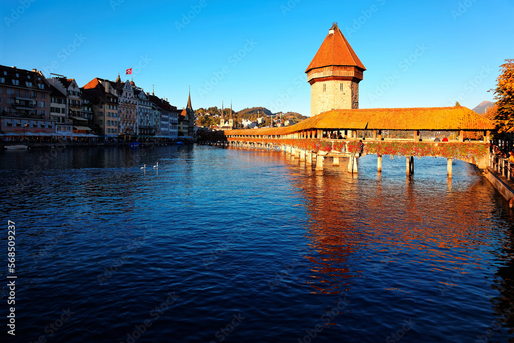 Scenery of Chapel Bridge ( Kapellbrucke ) and Water Tower bathed in warm golden sunlight over Reuss River in Lucerne Old Town, Switzerland, with beautiful reflections on the water under blue clear sky