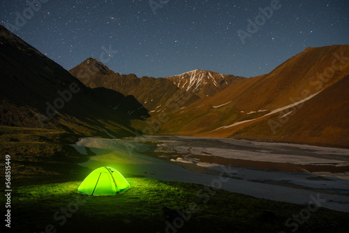 Night illuminated camping tent at frozen highland lake among dramatic mountain rocks with snow Altai Siberia Russia