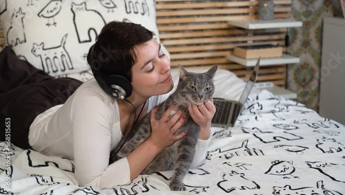 Beautiful woman is relaxing on the bed with her cat. The woman is playing with the gray cat while she listens to music.
