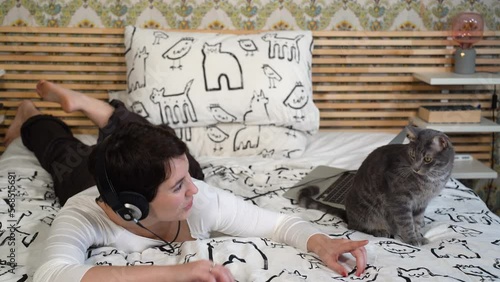 Beautiful woman is relaxing on the bed with her cat. The woman is playing with the gray cat while she listens to music.