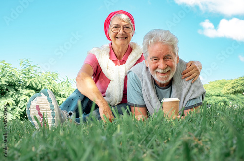 Lovely senior smiling couple lying on the grass in public park looking at camera. Romantic couple enjoying free time and retirement in nature #568530048