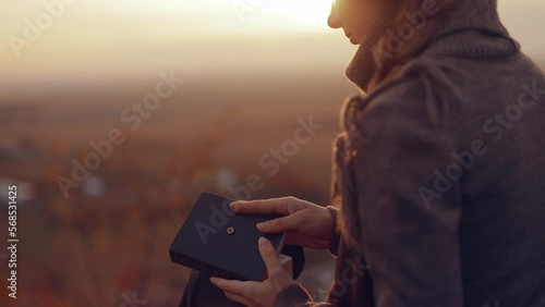 Close up Young Christian woman reading the bible, praying, putting her hand on the bible, sitting on a stone in sunset in autumn evening, peace and tranquility, faith, God's blessing, book of life. 