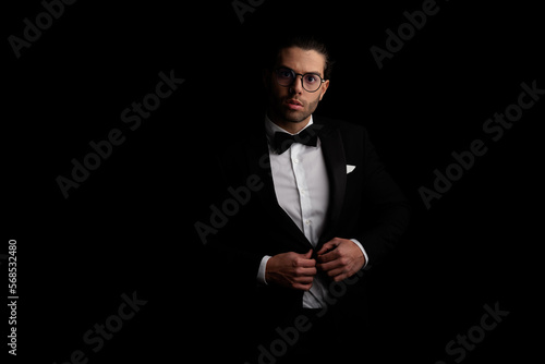 portrait of sexy arabic man with glasses adjusting and buttoning black tuxedo