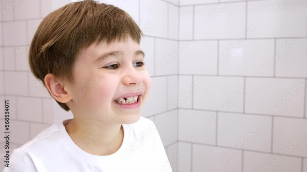 A child looks in the mirror at his loose milk tooth and pushes it with his tongue. Handsome caucasian 6 year old boy in a white T-shirt on a white bathroom background. Unhappy grimace. lower incisor. A child looks in the mirror at his loose milk tooth and pushes it with his tongue. Handsome caucasian 6 year old boy in a white T-shirt on a white bathroom background. Unhappy grimace. lower incisor.