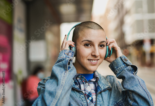 Young bald girl listening music playlist while waiting at bus station in the city