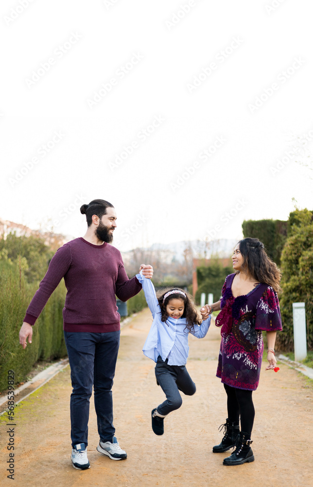 Obraz premium The interracial family is active and enjoys a day at the park. Successful adoption. The parents swing the daughter with their arms while they watch. Concept of multiethnic or interracial family.