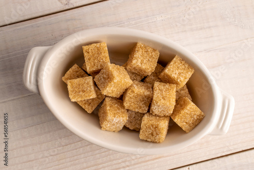 Several brown sugar cubes in a white ceramic bowl, close-up, on a white wooden table.