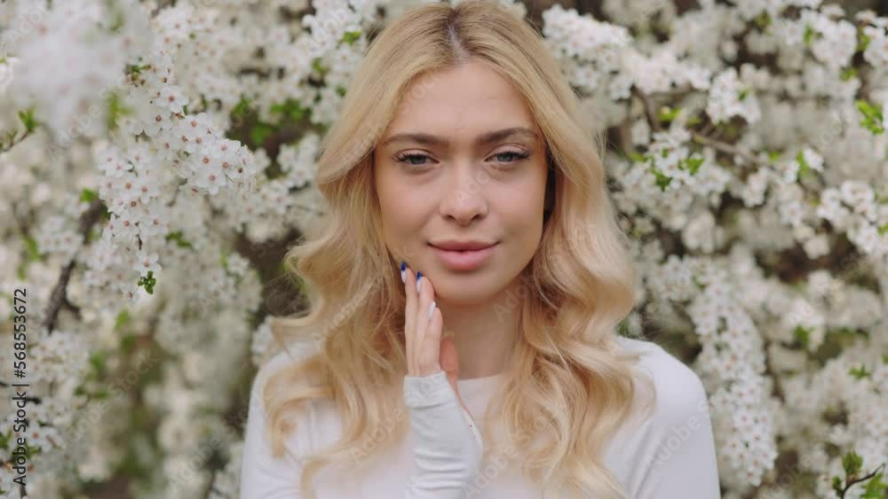 Gorgeous caucasian woman with wavy blond hair looking at camera while standing at spring orchard with beautiful seasonal beauty. White blossoming on trees around. Gorgeous caucasian woman with wavy blond hair looking at camera while standing at spring orchard with beautiful seasonal beauty. White blossoming on trees around.