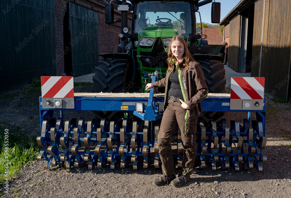 Obraz premium Stolz steht eine junge Landwirtin vor ihren Trecker mit einen landwirtschaftlichen Gerät angekuppelt, auf ihren Hof.