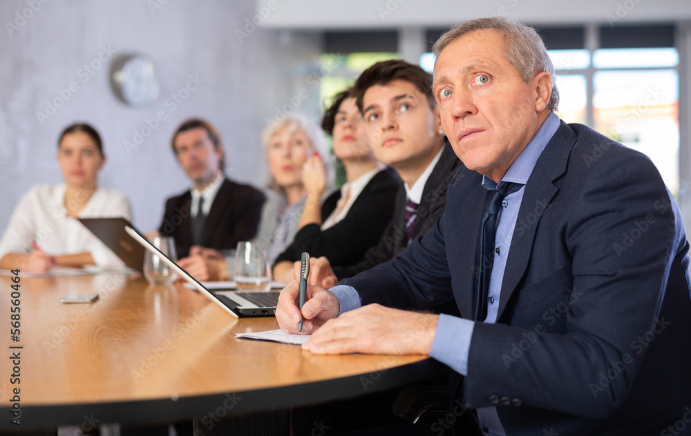 Fototapeta premium Portrait of surprised elderly businessman sitting at table with colleagues in office and watching presentation during business meeting