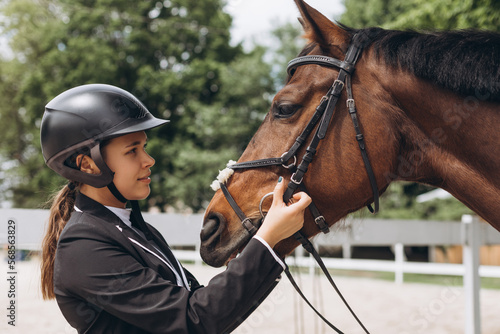Beautiful professional female jockey standing near horse. Woman horse rider is preparing to equitation