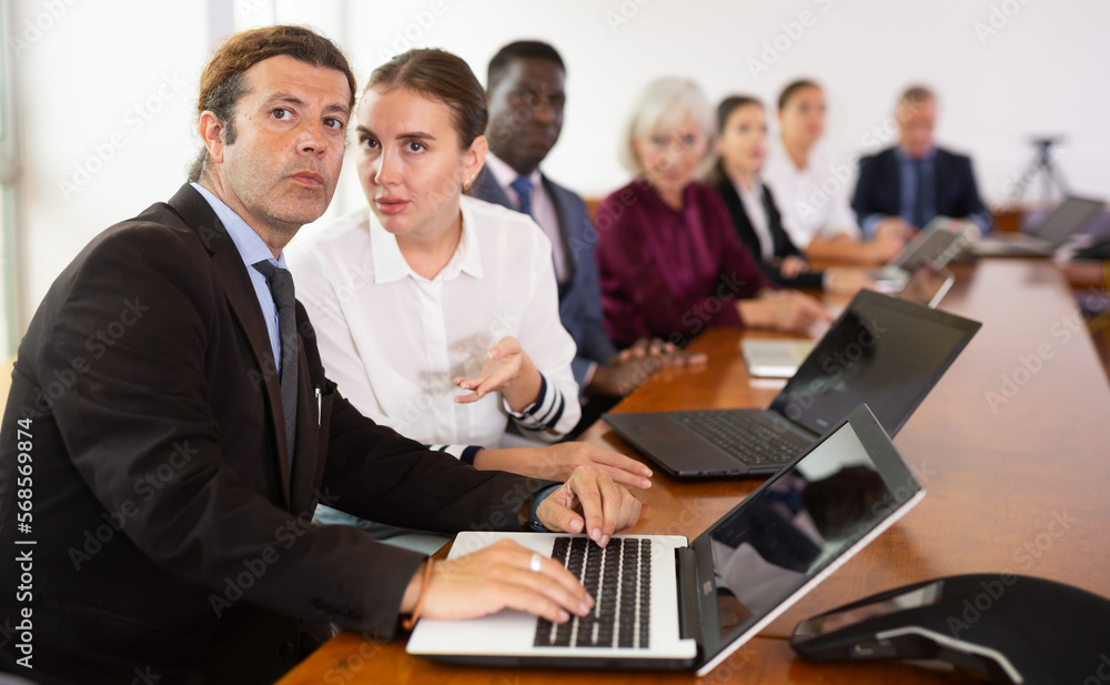 Fototapeta premium Portrait of middle aged white businessman and young white businesswoman sitting at table, working together using laptops and discussing business project at multiracial corporate group briefing in