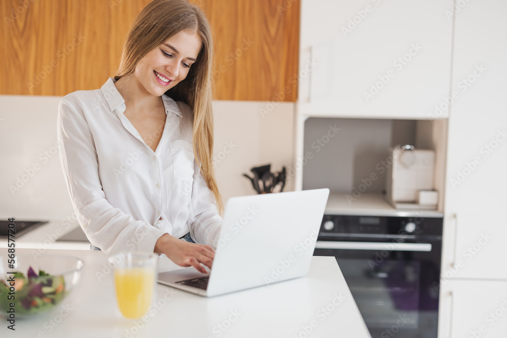 Happy and smiling blonde woman looking for recipe on laptop on internet in kitchen Happy and smiling blonde woman looking for recipe on laptop on internet in kitchen