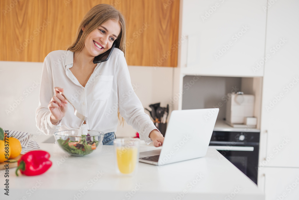Happy and smiling blonde woman talking on the phone and looking for a recipe on the internet on a laptop in the kitchen
