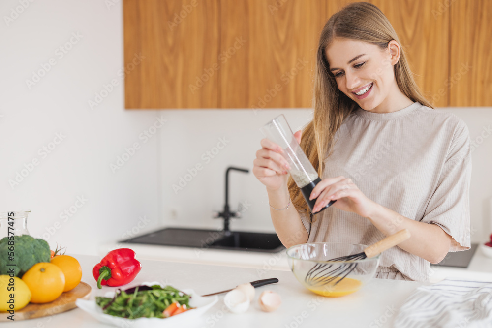 Happy pretty blonde woman in pajamas is preparing breakfast of eggs and salt it among fresh vegetables in the kitchen at home. Happy pretty blonde woman in pajamas is preparing breakfast of eggs and salt it among fresh vegetables in the kitchen at home.