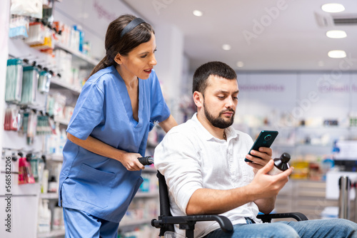 Female pharmacist consulting disabled man about medicine. Man sitting on wheelchair and using smartphone.