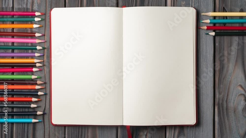 Top view of open empty blank notebook on grey wooden table. Different colored pencils appearing adding up in row. Back to school, art, drawing, stationary, office supplies. Flat lay, stop motion.