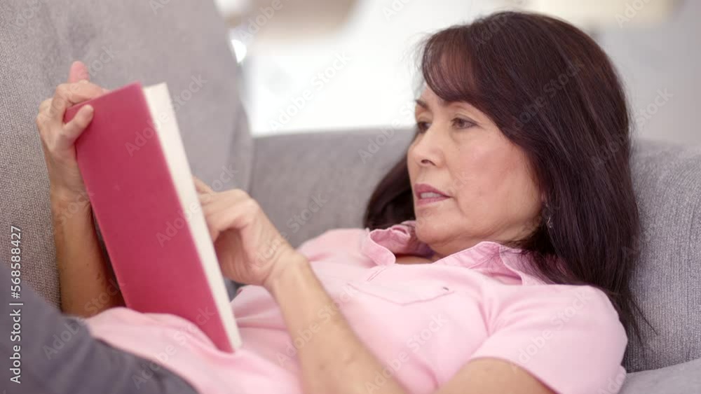 Attractive mature asian woman in pink blouse relaxing reading book laying on comfortable couch in leisure home living room Attractive mature asian woman in pink blouse relaxing reading book laying on comfortable couch in leisure home living room