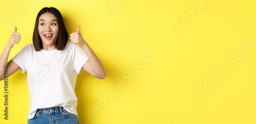 Fotografia Pretty young asian woman in white t-shirt, showing thumbs up and smiling, praise