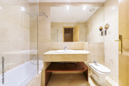 Bathroom with cream-colored marble furniture with an elongated bathtub and a mirror embedded in the wall and a metal shelf inside and a glass partition