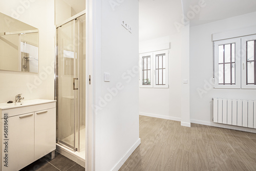 empty room with a pair of white aluminum windows with metal bars and access to a bathroom with white furniture and a shower cabin
