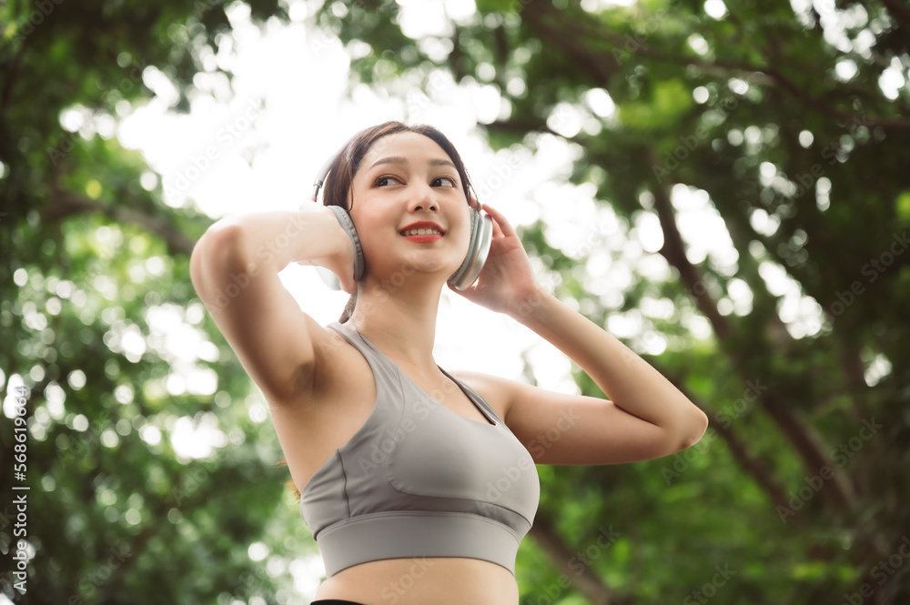 Asian woman trainer is engaged in fitness in public park. Female wearing sport bra and using headphone listening to music. Healthy lifestyle, sports outdoor activities in park