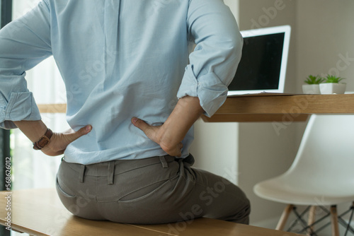 Working man sitting on the wood bench chair with laptop on desk in the office room. He used both hands to press down on the lower back. He is suffering from back pain from sitting for a long time.
