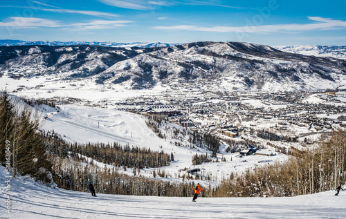 View of Colorado, USA, ski slope descending to ski resort village in winter with three figures of snowboarders going down the slope; distant mountains and blue sky in background