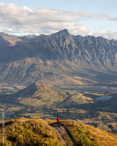 A young hiker stands at the top of a hill near Queenstown New Zealand, witnessing the beauty and awe of The Remarkables at sunset. (ID: 568630660)