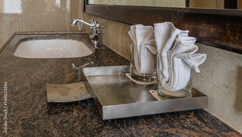 On the marble countertop in the bathroom there are glasses with folded napkins, a stainless steel soap dish. In the distance - a sink, a faucet. A mirror on a beige tiled wall. photo