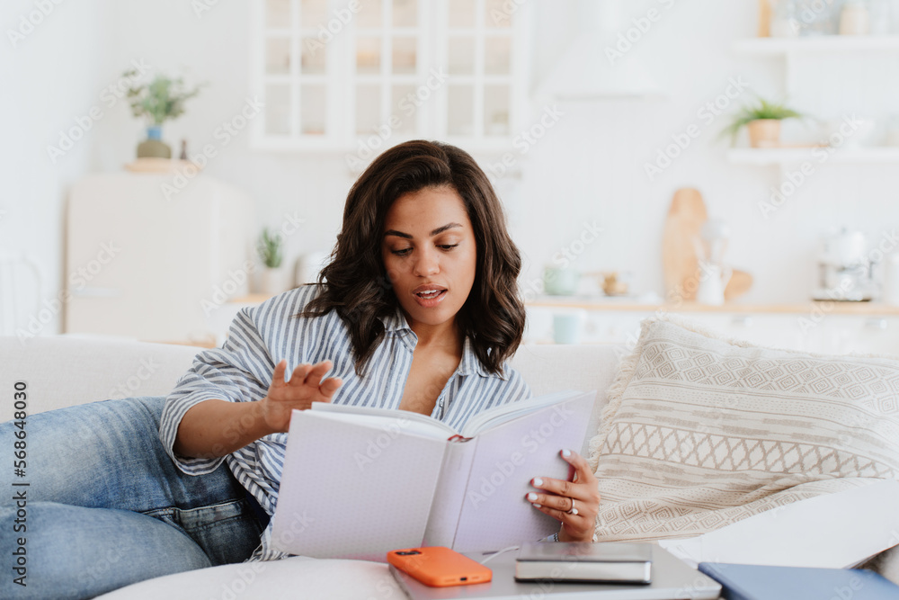 Beautiful African American girl in casual laying on cozy couch reads book having break of remote work. Brazilian businesswoman explores catalogue of furniture. Young woman resting on vacation. Beautiful African American girl in casual laying on cozy couch reads book having break of remote work. Brazilian businesswoman explores catalogue of furniture. Young woman resting on vacation.