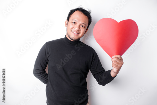 A portrait of a happy Asian man wearing a black shirt, holding a red heart-shaped paper isolated by white background © Reezky