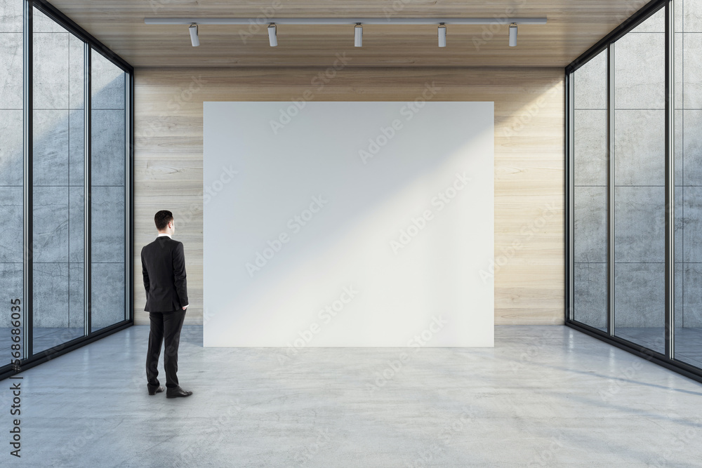 Man in black suit back view looking at blank white partition with place for your logo or advertising text on light wooden background in empty hall with transparent walls and concrete floor, mock up