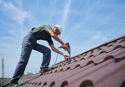 Wallpaper Mural Worker prepearing to installation photovoltaic solar panel system on the roof of house. Man installer working with electric screwdriver, wearing uniform and helmet, on blue sky background. Torontodigital.ca